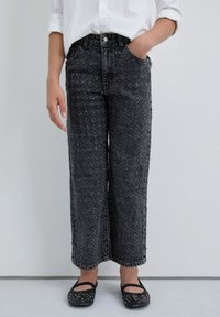 Jeans de mezclilla negra de pierna ancha con patrón texturizado, cinco bolsillos y cierre de botón. Combinados con bailarinas negras adornadas.