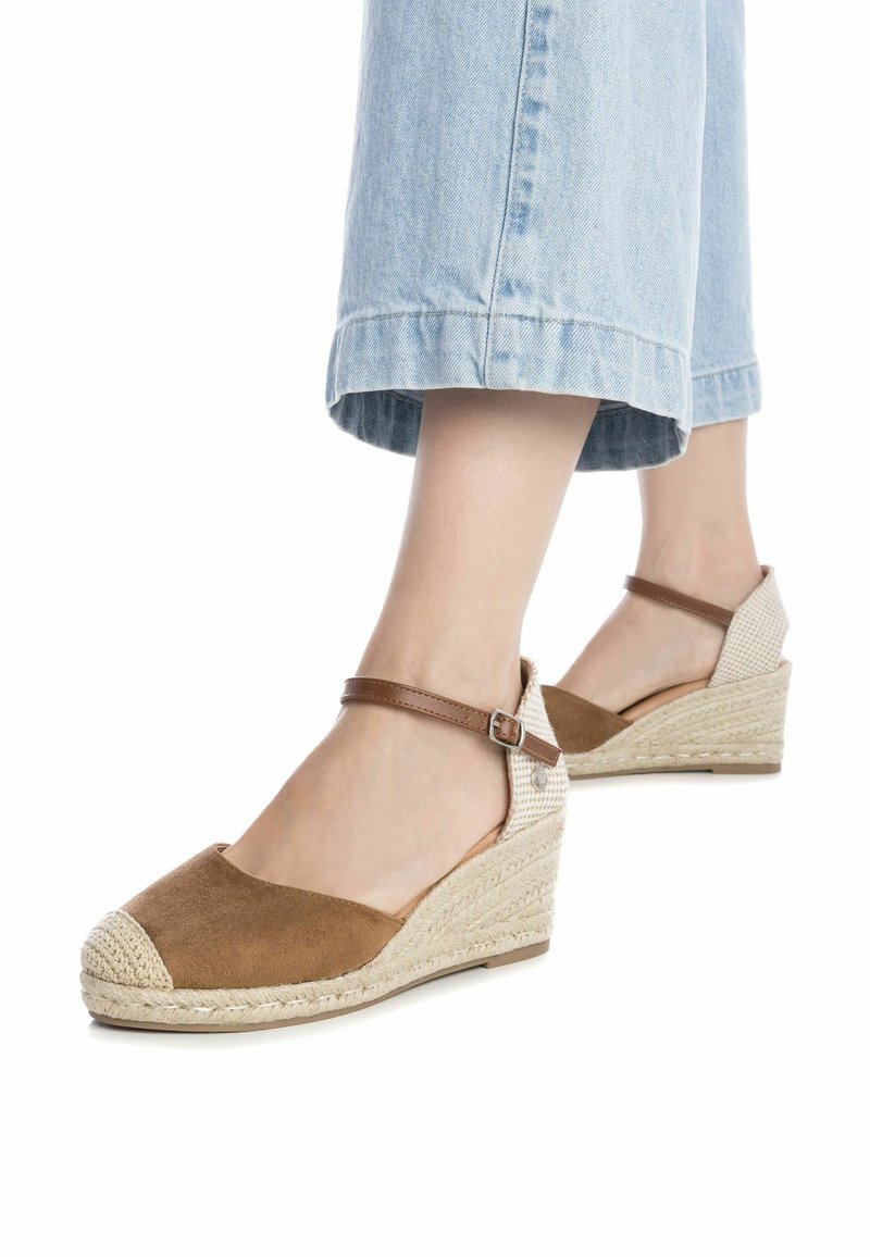 Pieds portant un jean bleu clair raccourci et des espadrilles compensées marron avec des brides à la cheville sur fond blanc.