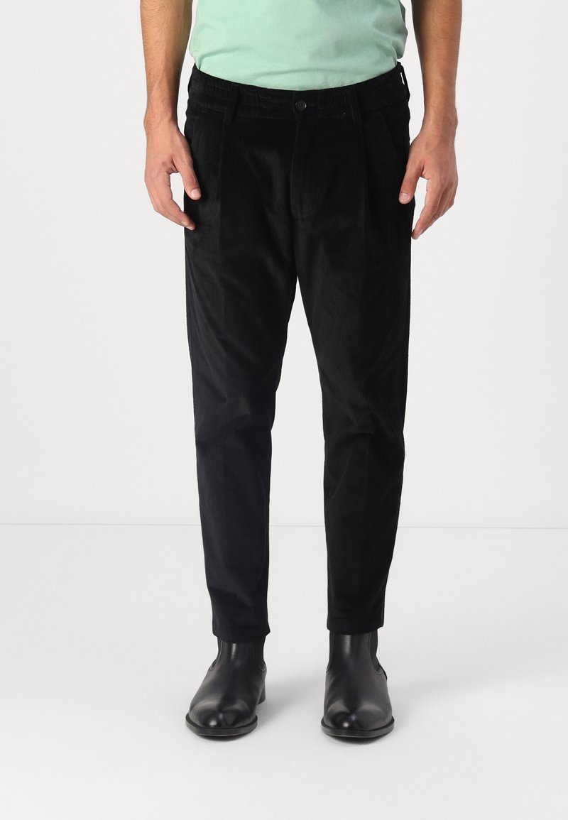 Homme portant un pantalon en velours côtelé noir, une chemise vert menthe et des bottines en cuir noir, debout devant un fond clair uni.