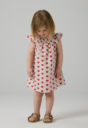 Lite barn i en hvit kjole med røde hjerte-mønstre som ser ned, iført beige sandaler, står foran en ensfarget grå bakgrunn.