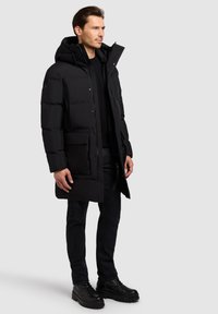 Giacca a vento nera con cappuccio, dotata di chiusura lampo frontale, tasche laterali e texture a coste. Abbinata a jeans scuri e stivali neri.
