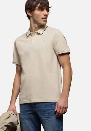 Homme portant un polo beige avec des bordures foncées et un jean bleu, tenant une veste beige assortie dans la main gauche, regardant vers sa droite.