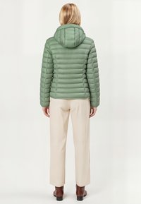 Lichtgroene pufferjas met een capuchon, voorzien van een horizontaal gestikt ontwerp. Gecombineerd met crèmekleurige broek en bruine schoenen, gezien van de achterkant.