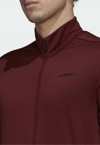 Chaqueta deportiva de color marrón burdeos con cremallera y un alto cuello, que presenta una textura sutil y un logotipo bordado en negro "Terrex" en el lado izquierdo del pecho.