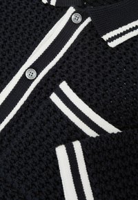 Black knit cardigan with textured pattern, white striped trim along the button line, cuffs, and pocket edges, and silver buttons.