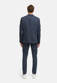 Mannen donkerblauw pak gemaakt van gestructureerde stof, met een getailleerde pasvorm en subtiel patroon. Gecombineerd met witte sneakers. Van achteren bekeken.