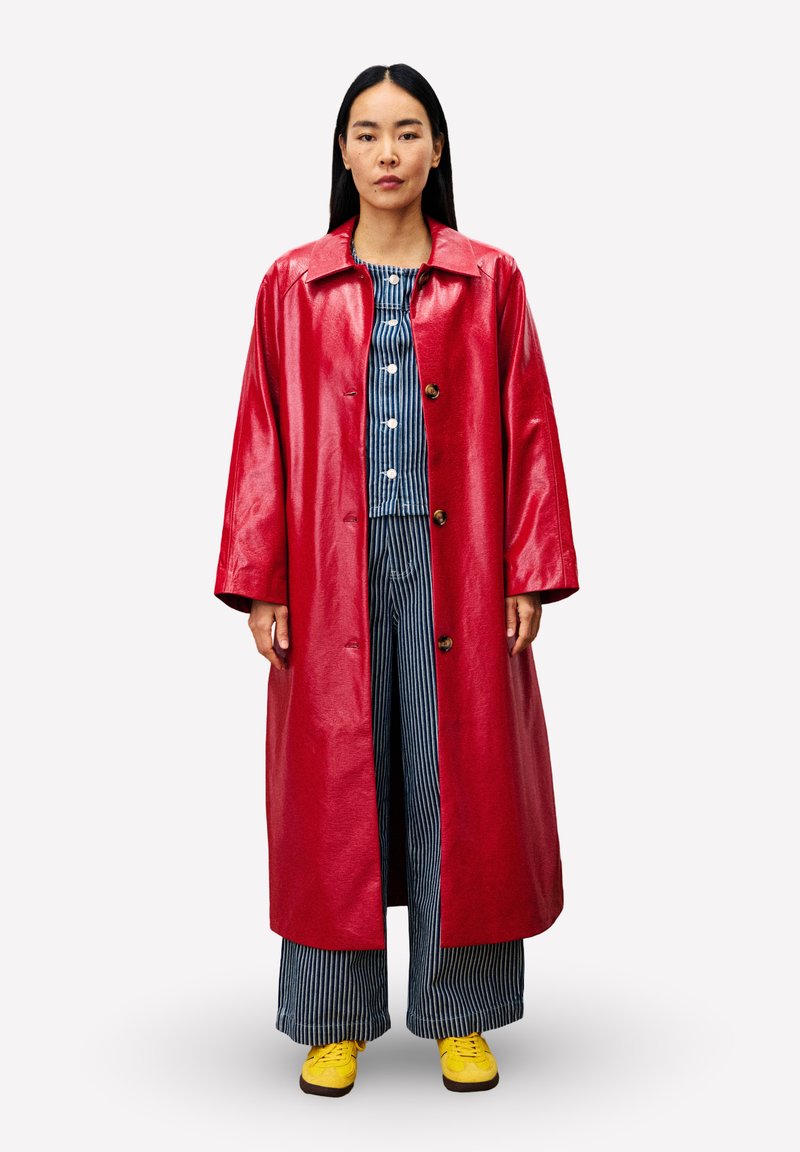 Frau mit langen, dunklen Haaren, die einen glänzenden roten langen Mantel über einem blau-weißen gestreiften Outfit und gelben Sneakers trägt, steht vor einem weißen Hintergrund.