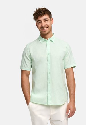 Uśmiechnięty mężczyzna z krótkimi kręconymi włosami, ubrany w jasnozieloną koszulę z krótkim rękawem na guziki i białe spodnie na jednolitym tle.