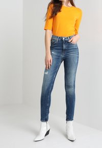 Top naranja de manga corta combinado con jeans azul de cintura alta con detalles desgastados; la modelo lleva botas blancas hasta el tobillo con tacón bloque.