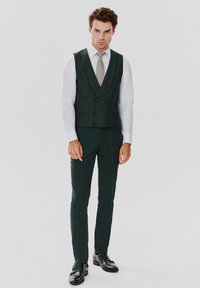 Ciemnozielona kamizelka z głębokim dekoltem w kształcie litery V, podwójnie zapinana, noszona na białej koszuli, w połączeniu z dopasowanymi spodniami i czarnymi butami wizytowymi.