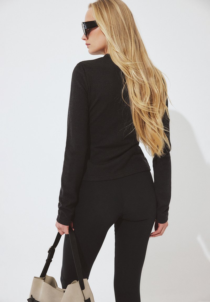 Naine pikkade blondide juuste, musta riietuse ja päikeseprillidega, hoides ühe käega beeživärvilist käekotti, näoga valge ja ühtlase tausta suunas.