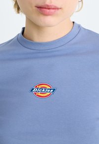 Világoskék pamut póló, amelyen egy piros, sárga és kék kör alakú logo található, középen a "Dickies" felirattal.