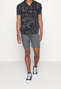 Man in een zwart shirt met witte abstracte gezichten, grijze shorts en zwarte hoge sneakers met witte veters, aan het lopen tegen een effen achtergrond.