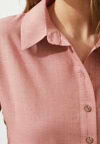 Chemise boutonnière à manches courtes de couleur rose pâle, dotée d'un col, de petits boutons et d'un tissu doux et texturé.