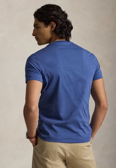 Sötét göndör hajú férfi kék rövid ujjú ingben és bézs nadrágban, kezét zsebében tartva, oldalra nézve, semleges háttér előtt áll.