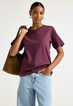 Kvinde iført en lilla kortærmet skjorte og lyseblå jeans, der bærer en brun ruskinds-totepose over skulderen.