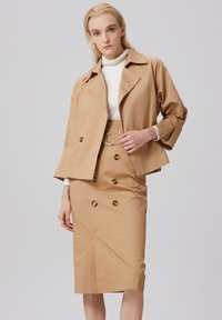 Ensemble composé d'une veste beige et d'une jupe crayon, fabriqué à partir d'un tissu lisse. La veste possède un col cranté et des boutons double boutonnage ; la jupe présente une taille ceinturée.