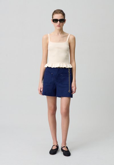 Débardeur beige à côtes avec ourlet volanté, associé à un short bleu marine avec poches et détail en chaîne. Des chaussures noires à motifs complètent le look.