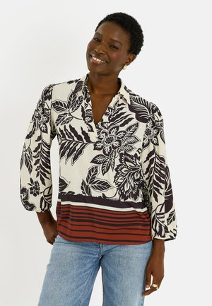 Mujer sonriente con una blusa floral en blanco y negro con dobladillo a rayas en color óxido y negro y vaqueros azul claro sobre un fondo liso.