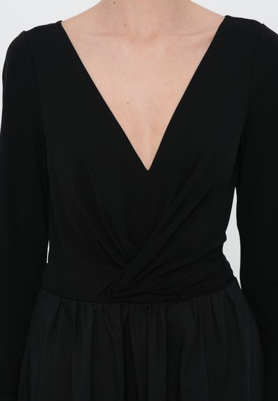 Juoda suknelė su ilgomis rankovėmis, gilus V formos iškirptė, suspausta liemeniu ir plačia sijonu. Pagaminta iš minkštos medžiagos su lygiu paviršiumi.