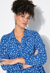 Blå blus med hjärtformade mönster i ljusblått, med krage, knappstängning fram och elastiska ärmslut; mjuk tygkvalitet.