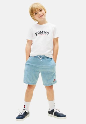 Blond dziecko stoi uśmiechnięte z rękami w kieszeniach, ubrane w białą koszulkę Tommy Hilfiger, jasnoniebieskie szorty, białe skarpetki i granatowe trampki.