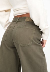 Pantalon large kaki avec deux poches arrière, détails cousus et étiquette de marque visible sur la ceinture. Tissu doux et texturé.