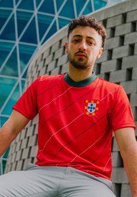 Rote Fußballtrikot mit grünem Kragen, das diagonale weiße Streifen und ein Wappen mit einem Wappen zeigt. Das Modell trägt graue Hosen.