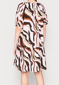 Mujer vestida con un vestido de longitud hasta la rodilla con rayas onduladas abstractas en marrón, beige, rosa, blanco y naranja, vista desde atrás.