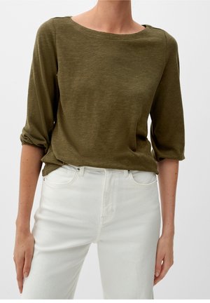 Femme portant un haut col bateau vert olive à manches 3/4 et un pantalon blanc taille haute, mains détendues le long du corps sur un fond uni.