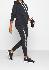 Svart huvtröja med vita snören, tillsammans med svarta joggers med "Under Armour" märkning i vitt och svarta träningsskor.