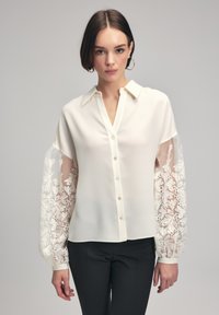 White button-up shirt with long, sheer embroidered sleeves featuring floral patterns. Soft fabric with a classic collar and front buttons.