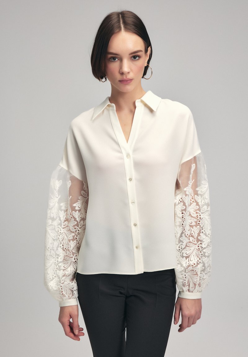White button-up shirt with long, sheer embroidered sleeves featuring floral patterns. Soft fabric with a classic collar and front buttons.