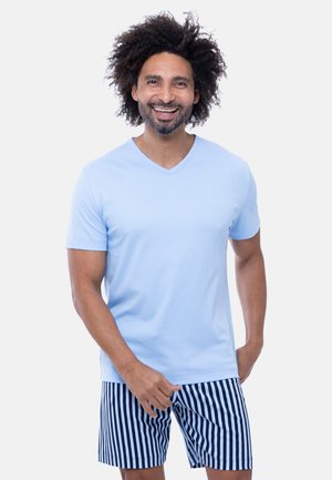 Lachende man met krullend haar draagt een lichtblauw, kort mouw shirt en marineblauw met witte verticale gestreepte korte broek op een witte achtergrond.