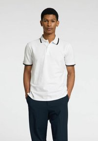 Polo shirt blanc avec col et bord des manches noirs, associé à un pantalon foncé. À manches courtes, doté d'une patte de boutonnage et d'une coupe décontractée.