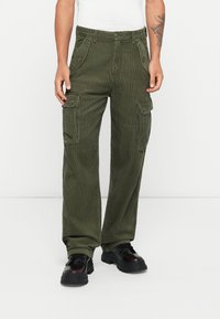 Pantalones cargo de pana verde olivo con múltiples bolsillos laterales, un corte relajado y textura de canalé visible. Combinados con zapatos negros con cordones.