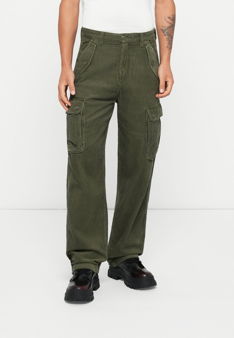 Pantalones cargo de pana verde olivo con múltiples bolsillos laterales, un corte relajado y textura de canalé visible. Combinados con zapatos negros con cordones.