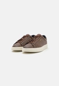 Barna bőr sneaker, sima felülettel, kerek fűzőkkel, fehér gumitalppal és diszkrét logózással az oldalon.