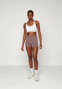 Biały stanik sportowy z cienkimi ramiączkami, w zestawieniu z brązowymi szortami sportowymi. Model nosi białe buty i białe skarpetki. Gładki, przylegający materiał.