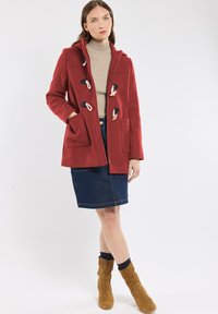 Manteau à capuche rouge avec des fermetures à bascule, deux poches avant, porté par-dessus un col roulé beige et une jupe en jean, associé à des bottines tan.