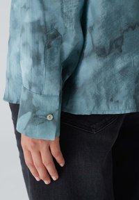 Camicia azzurro chiaro con una texture acquerellata, polsini abbottonati e jeans scuri a contrasto. Presenta una finitura liscia e una vestibilità sartoriale.