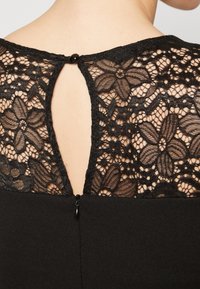 Svart klänning med en blommig spetsrygg som har en nyckelhålsöppning och en liten knappknäppning, samt en dold dragkedja vid den nedre delen av ryggen.