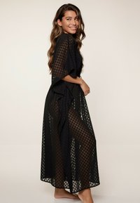 Capa tipo kimono negra y transparente con patrón geométrico, mangas anchas y un diseño fluido que revela las piernas debajo.