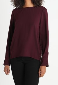 Blouse bordeaux à manches longues avec un col rond, fabriquée en tissu lisse. Dotée de manches ajustées et d'un ourlet légèrement arrondi. Portée avec un pantalon noir.