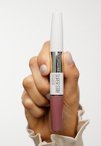 Rouge à lèvres liquide Maybelline Super Stay dans un tube transparent, avec un capuchon blanc, une teinte rose mate, et une étiquette argentée indiquant le nom du produit.