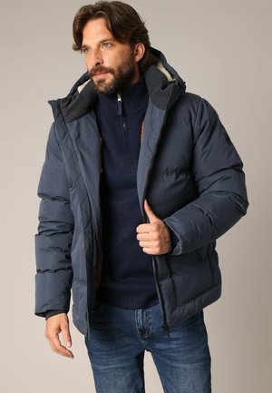 Doudoune marine avec capuche, présentant un design matelassé, une fermeture éclair et des accents marron. Superposée à un pull en maille marine.