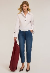 Camicetta bianca con colletto e bottoni decorativi, abbinata a pantaloni in denim blu. Tiene in mano un blazer rosso e indossa tacchi neri lucidi.