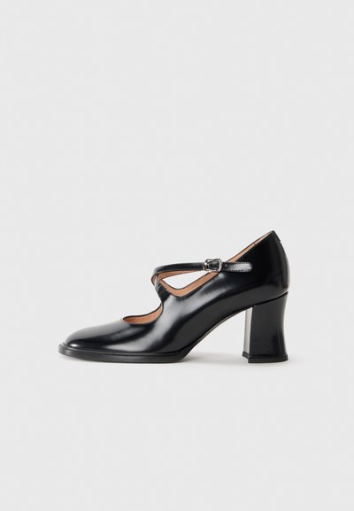 Des chaussures à talons hauts en cuir verni noir avec un design découpé, présentant une bride de cheville et un talon épais. Texture lisse et bout arrondi.