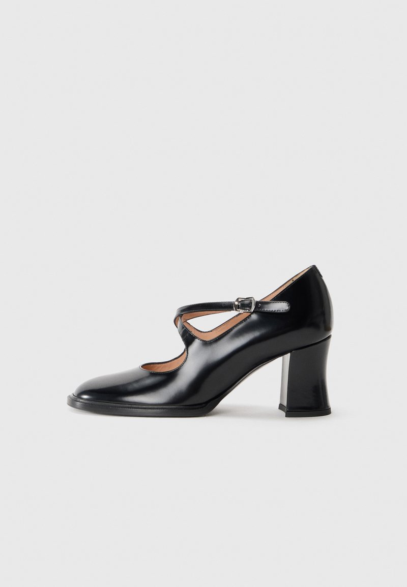 Des chaussures à talons hauts en cuir verni noir avec un design découpé, présentant une bride de cheville et un talon épais. Texture lisse et bout arrondi.