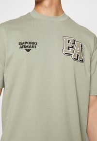 Jasnozielona bawełniana koszulka z krótkim rękawem, z czarnym logo "Emporio Armani" oraz beżowym haftem "EA" na lewej piersi.